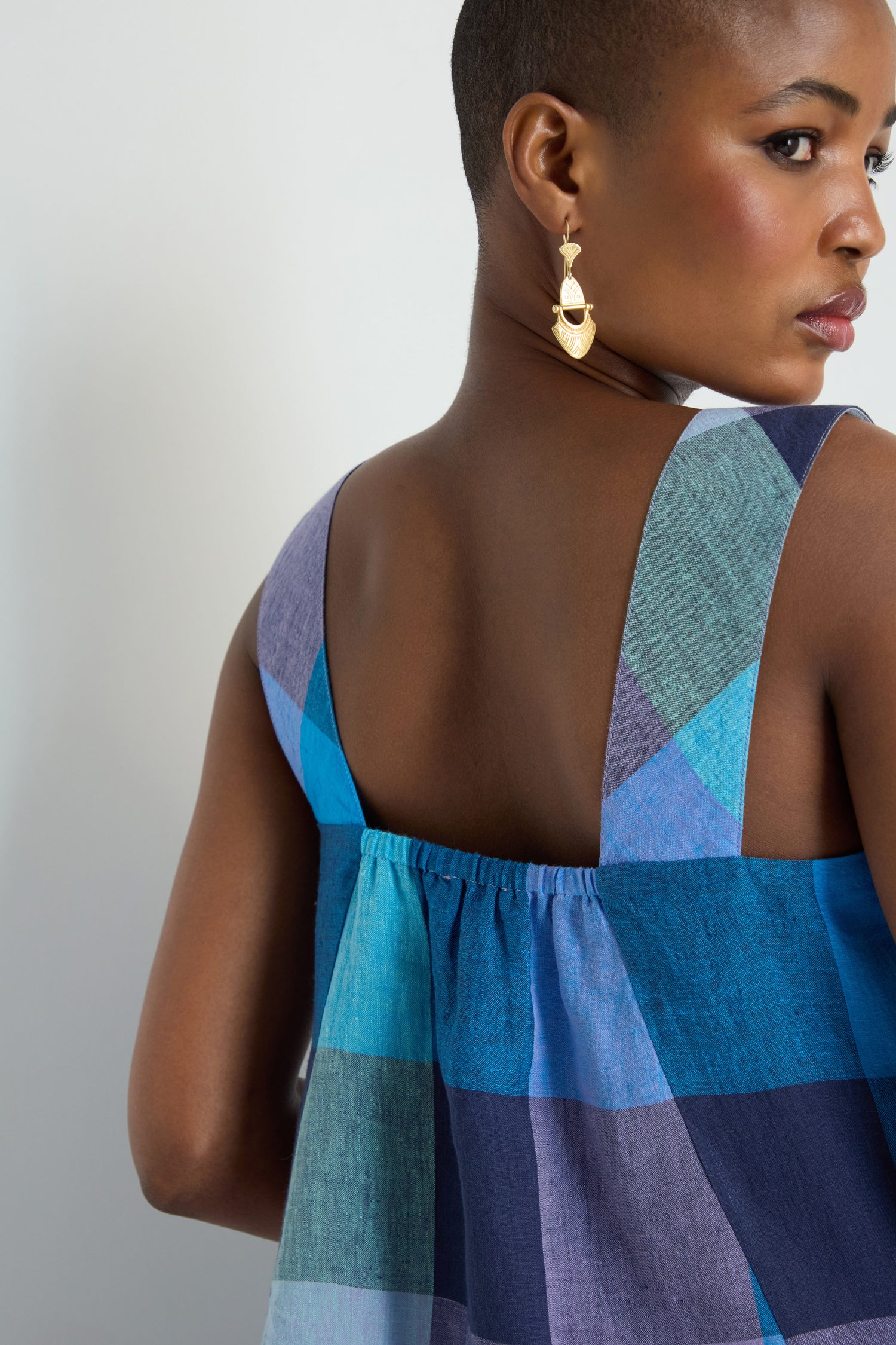 A person with short hair and gold earrings models the Cross Dye Check Linen Bubble Dress—sleeveless, blue plaid linen—in a back view, turning slightly toward the camera.