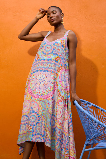 A woman wearing a colorful Circle Mandala Sleeveless Dress stands against an orange wall, gently touching her head with one hand while the other rests on a blue chair.