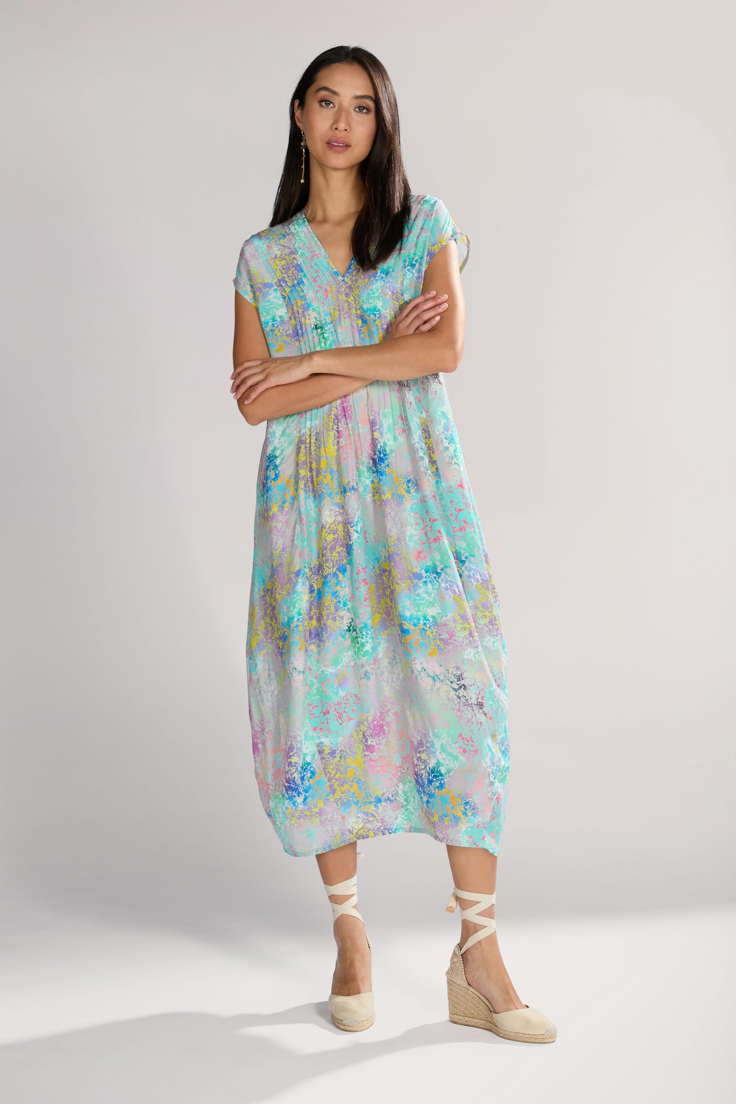 A woman stands with her arms crossed, wearing a pastel-colored, floral-patterned long dress and beige wedge sandals. The Marbled Cloud Print Bubble Dress gives an elegant and eye-catching touch to her ensemble.