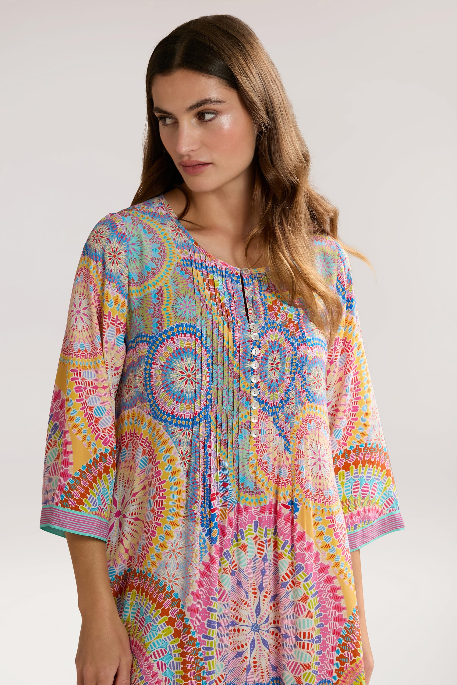 A woman with long brown hair is wearing a colorful Circle Mandala Pleat Front Dress with intricate designs. She is looking slightly to her left against a plain background.