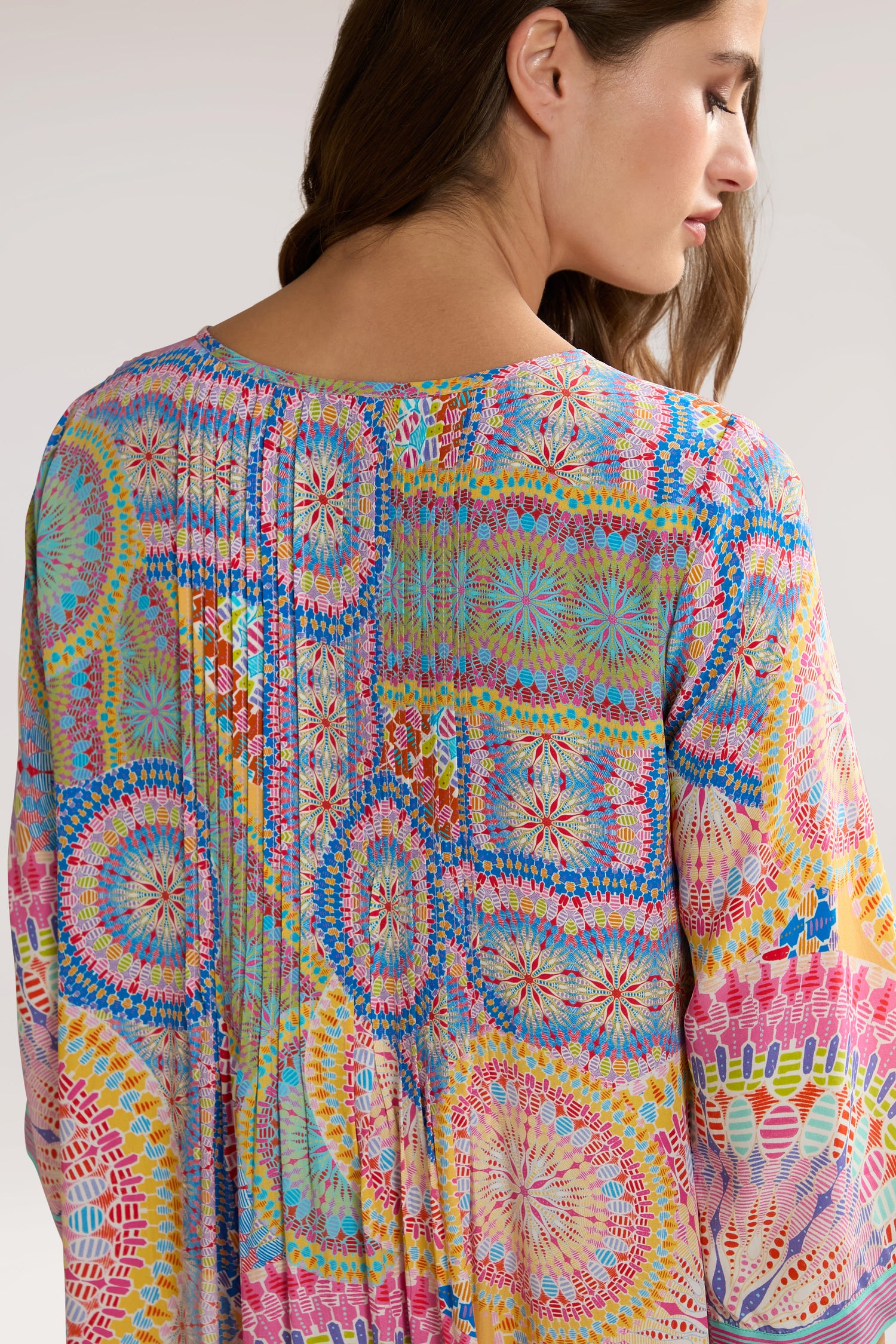 A person wearing a Circle Mandala Pleat Front Dress, shown from the back. The fabric features an array of vibrant, circular and geometric designs reminiscent of a mandala print.