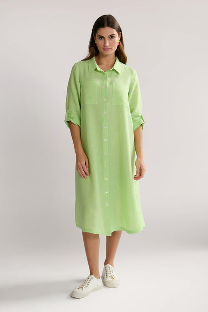 A woman stands wearing a light green, button-up, long-sleeve dress made from natural fibres. She pairs the effortlessly chic Linen Ticking Stripe Shirt Dress with white sneakers.