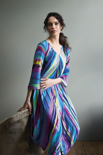 A woman wearing the Watercolour Landscape Bubble Dress, characterized by its vibrant and striped design made from lightweight viscose fabric, has her hair tied back as she leans against a cushioned surface and looks towards the camera.