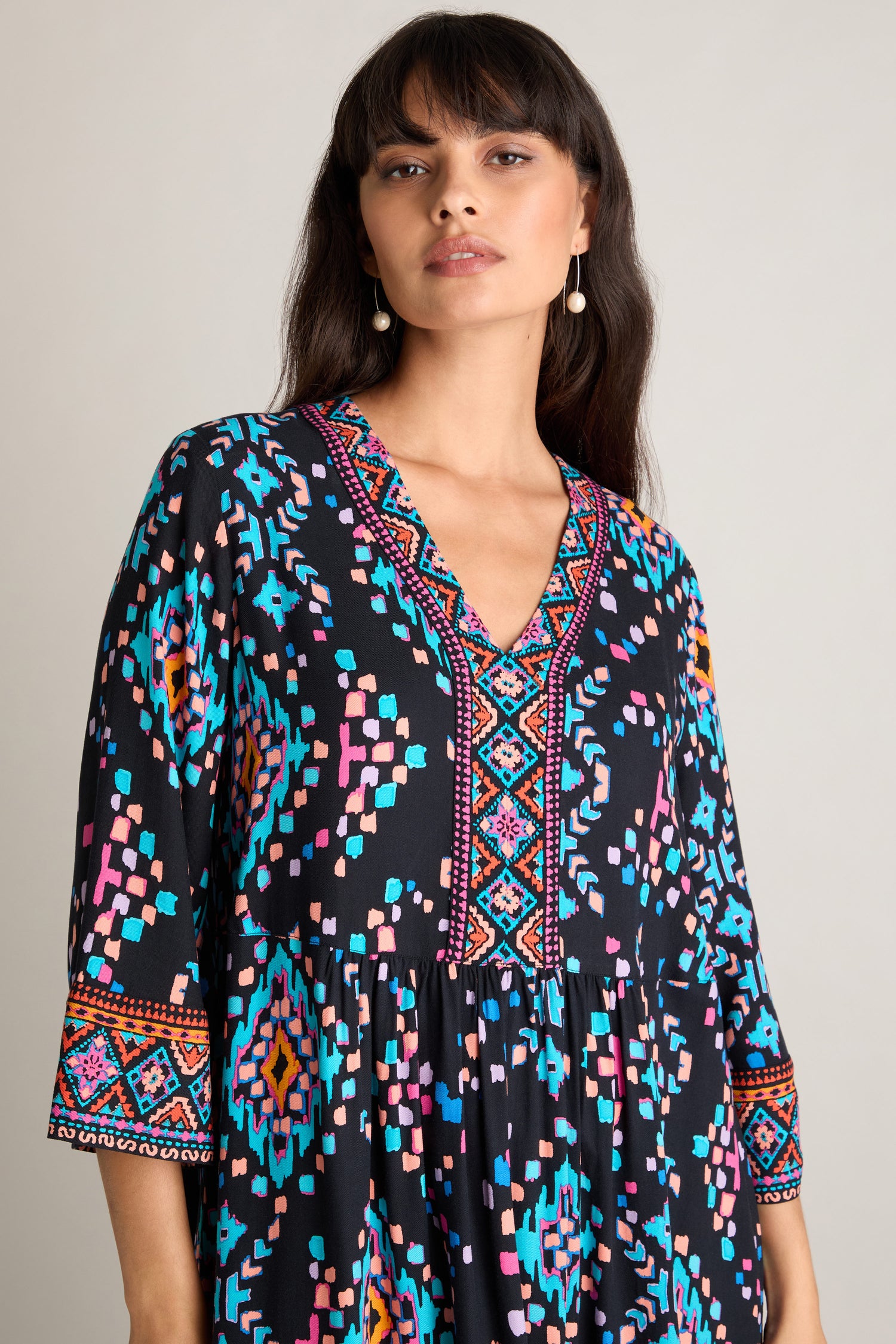 A woman is wearing the Geo Kilim Dress in black, which showcases a vibrant and colorful geometric pattern. The dress features a deep V-neckline and three-quarter sleeves adorned with patterned detailing. She has long hair and is accessorized with earrings.