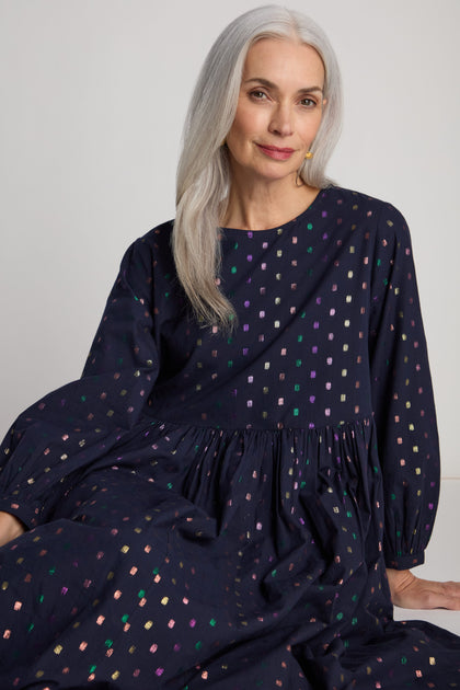 A woman with long gray hair is seated against a neutral background, looking at the camera with a slight smile. She is wearing a dark Multi Square Lurex Pleated Dress adorned with colorful sparkle details.