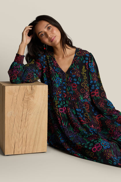 A woman in an Embroidered Floral Print Dress sits on the floor, resting her elbow on a wooden block and looking at the camera.