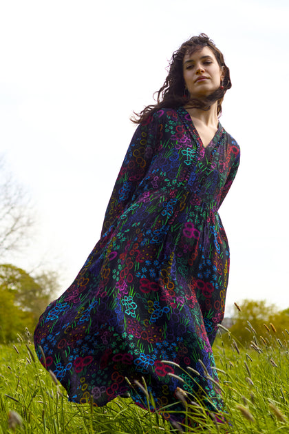 A person wearing a Floral Embroidery Print Dress, featuring a long, black V-neck design with delicate craft floral patterns, stands in a grassy field under a bright sky.