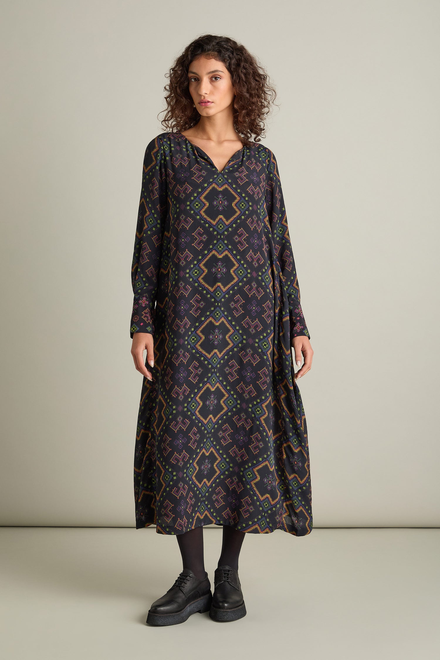 A woman with curly hair stands against a plain background, wearing a long Pixelated Jacquard Print Dress in silky crepe viscose, accompanied by black leggings and shoes.