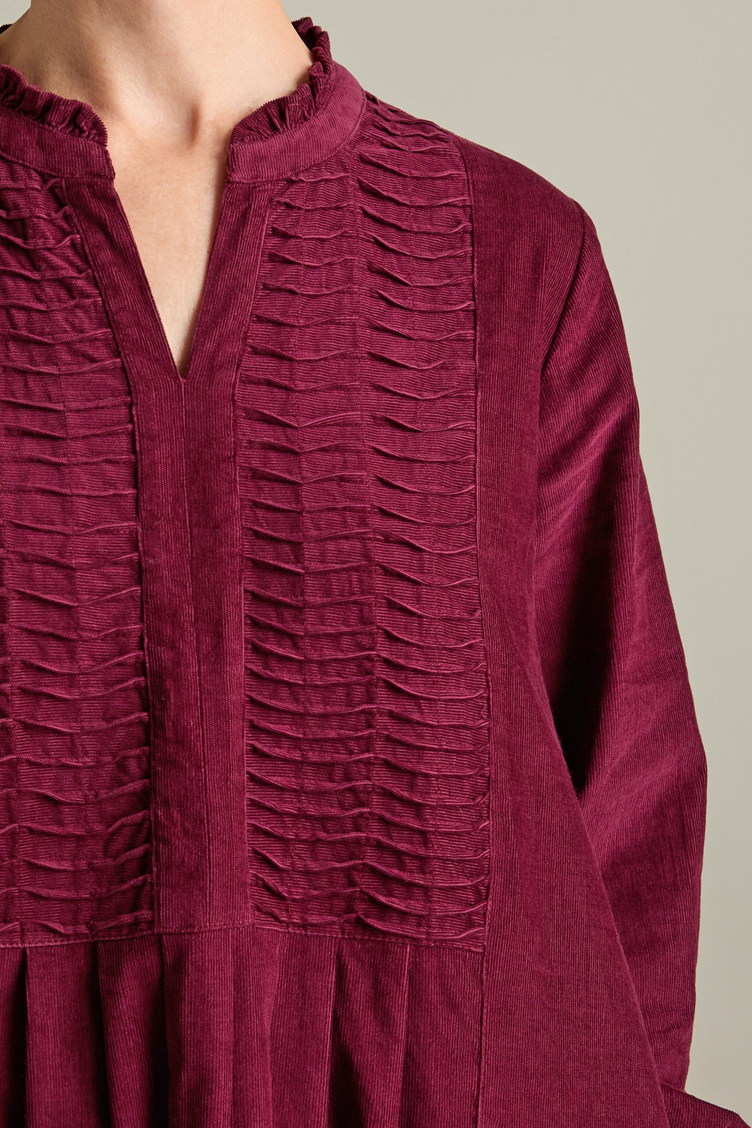 Close-up of a person wearing the Pinwale Cord Pleat Front Dress, showcasing its maroon textured fabric with vertical pleats, a v-neckline, and multi-pin tucking yoke detailing.