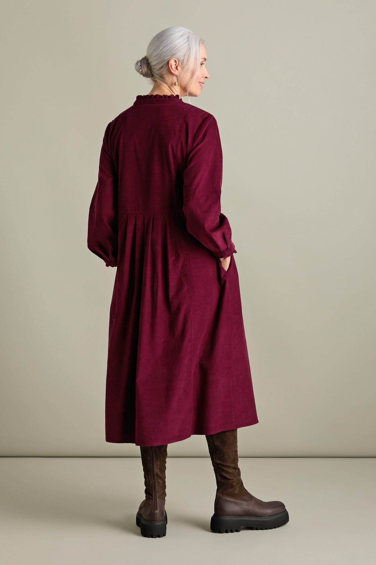 Woman with gray hair in a maroon Pinwale Cord Pleat Front Dress featuring a Multi-pin Tucking Yoke and an A-line Silhouette stands looking to the side, brown boots completing her elegant look.