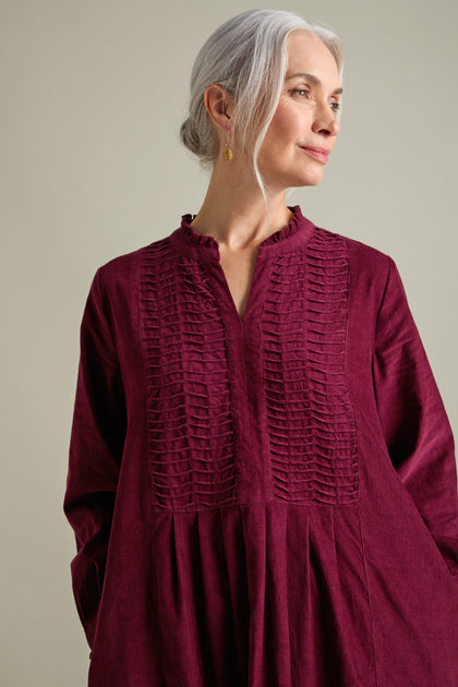 An older woman with long gray hair, adorned in a deep red textured Pinwale Cord Pleat Front Dress featuring a high collar, gazes sideways. The A-line silhouette enhances her elegant stance against the plain, light-colored background.