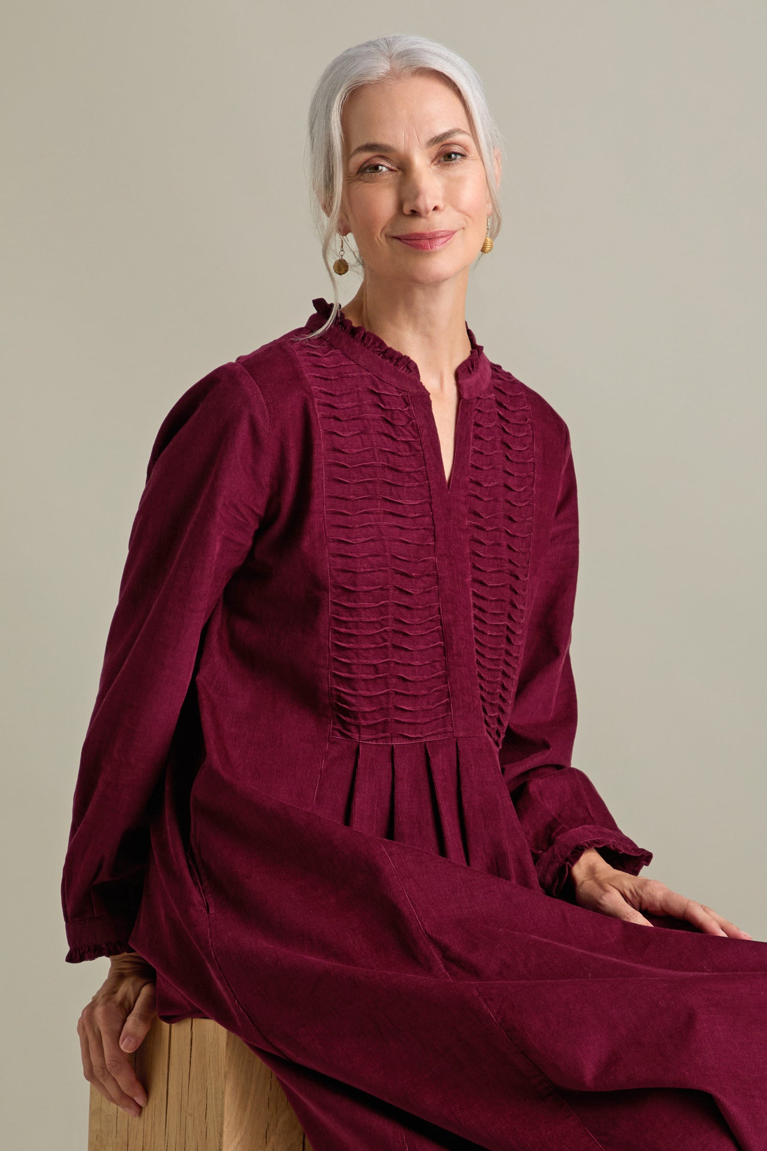 A woman with long, gray hair is seated on a wooden stool, smiling slightly. She is wearing the Pinwale Cord Pleat Front Dress in burgundy, which showcases a multi-pin tucking yoke and an elegant A-line silhouette. The background is plain and light-colored.