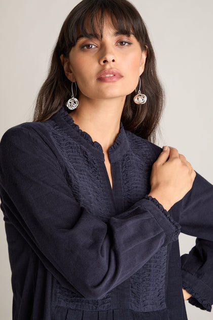 A person with long, dark hair is wearing a Pinwale Cord Pleat Front Dress in navy blue with detailed stitching and silver spiral earrings, posed with one arm bent across the chest, showcasing an elegant A-Line silhouette.