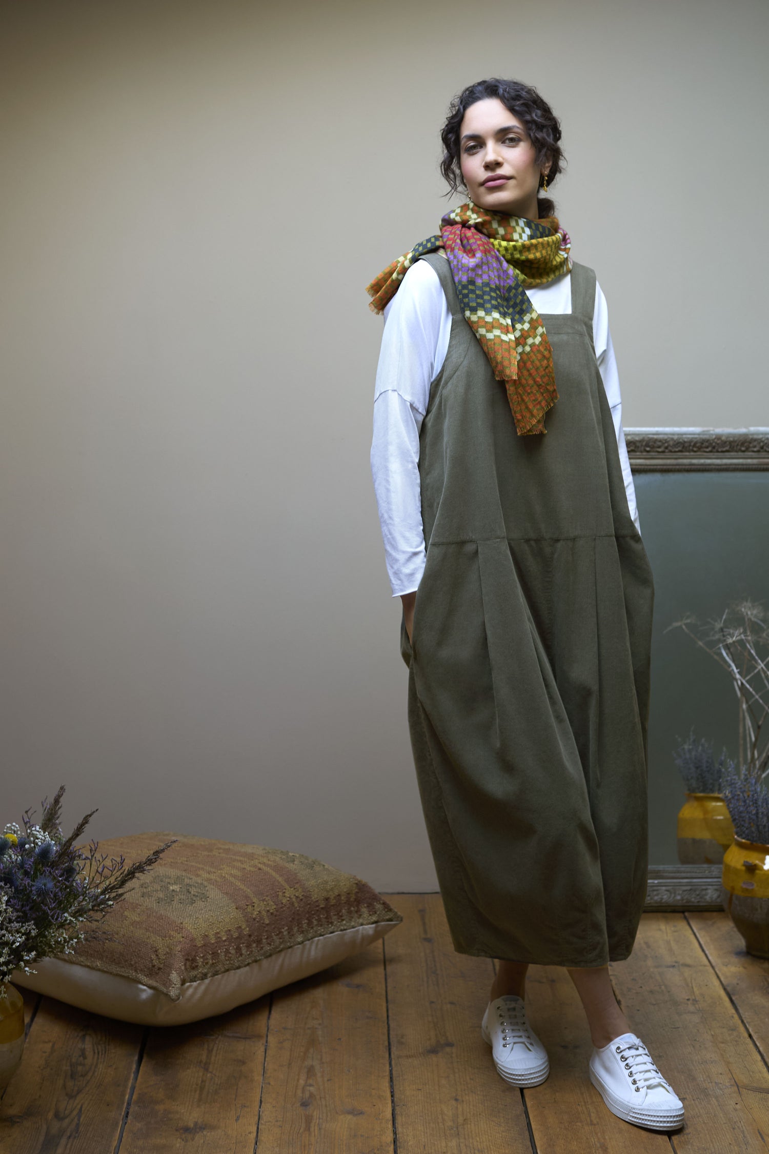 A person wearing a white long-sleeved shirt under a green Pinwale Cord Jumpsuit, accessorized with a colorful scarf, white shoes, standing on a wooden floor next to a patterned cushion and framed artwork.