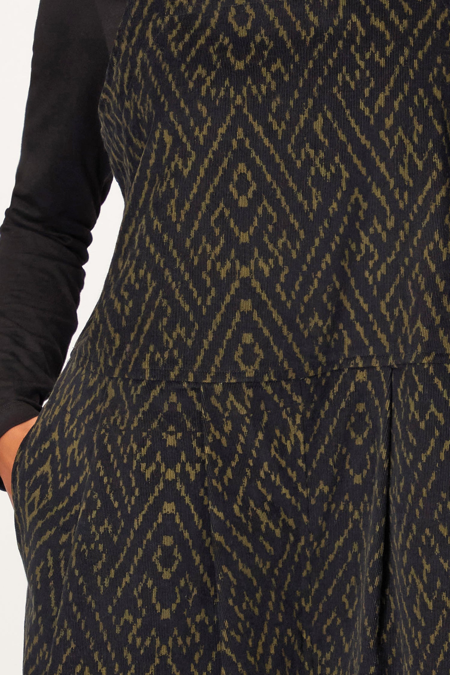 A person wears the Ornate Textured Cord Jumpsuit, a dark, textured statement piece with relaxed tailoring and pockets, paired with a black long-sleeve top—shown from shoulders to mid-thigh.