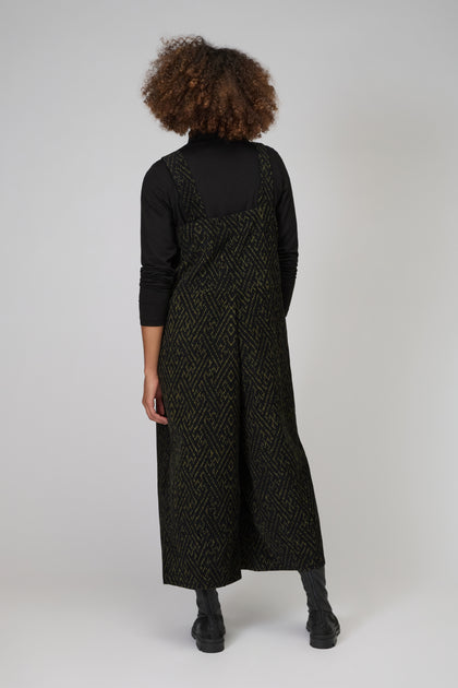 Person facing away, wearing a statement piece: the Ornate Textured Cord Jumpsuit over a black turtleneck and boots, standing against a plain light background.