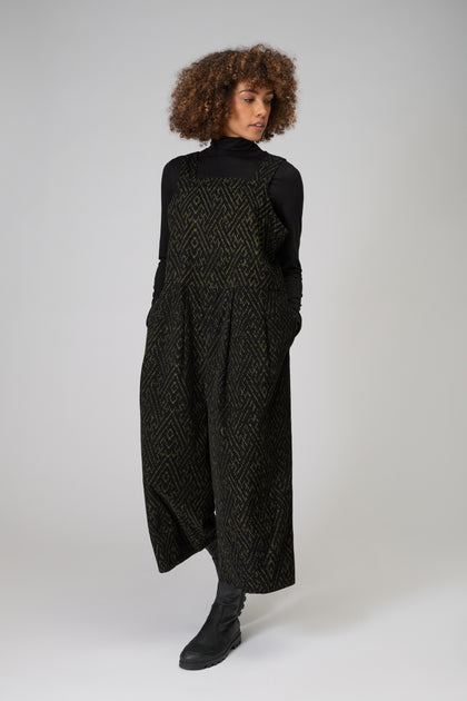 A person with curly hair wears a black turtleneck and the Ornate Textured Cord Jumpsuit with pockets, styled with black boots, standing against a plain light background.