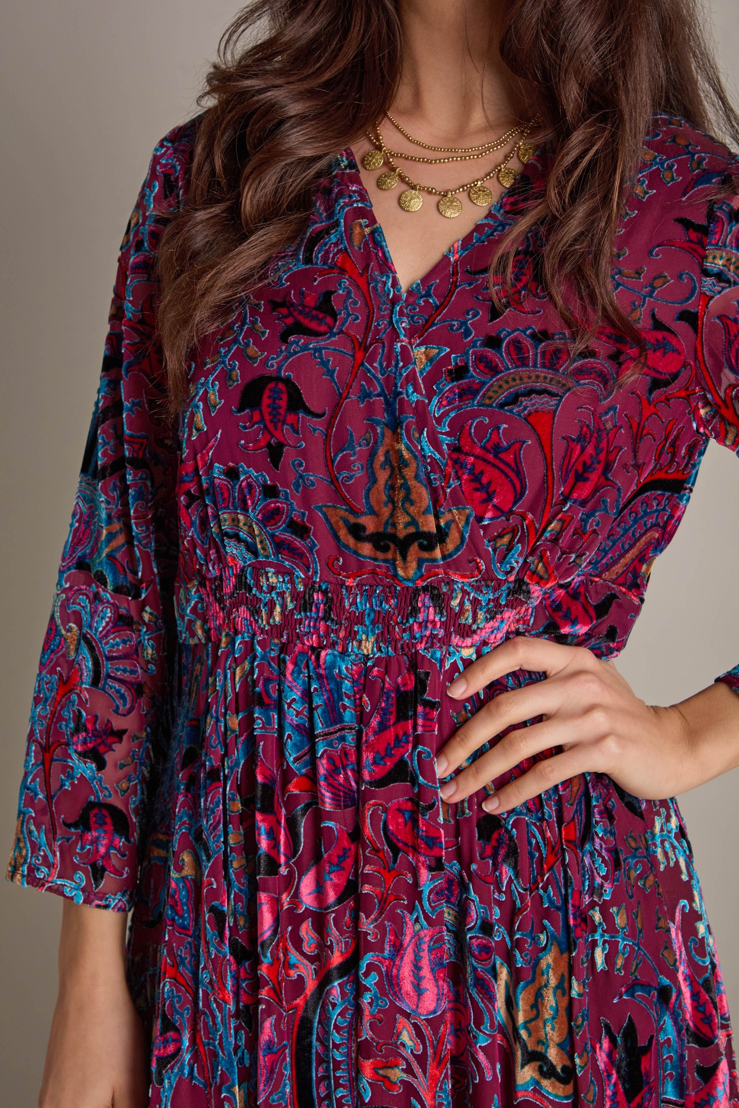 A person confidently poses with one hand on their hip, wearing the Winter Paisley Devore Flared Dress with an A-line silhouette and long sleeves, adorned with gold necklaces.