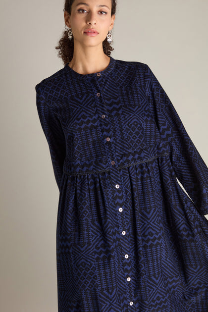 A person wearing a long-sleeved, button-up Mono Ikat Printed Cord Dress featuring a blue and black geometric patchwork print stands against a plain background.