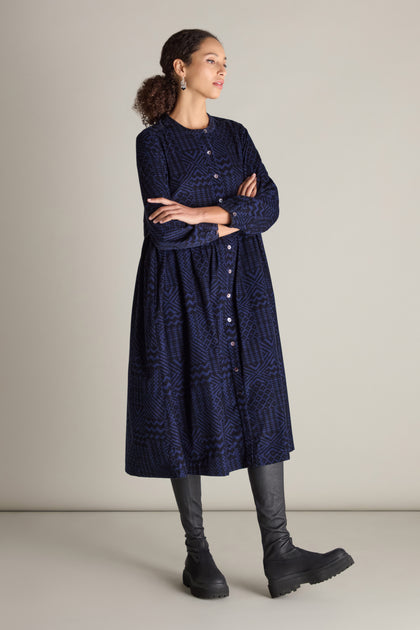 A woman stands with her arms crossed against a plain background, wearing a long-sleeved dark blue Mono Ikat Printed Cord Dress and black boots.