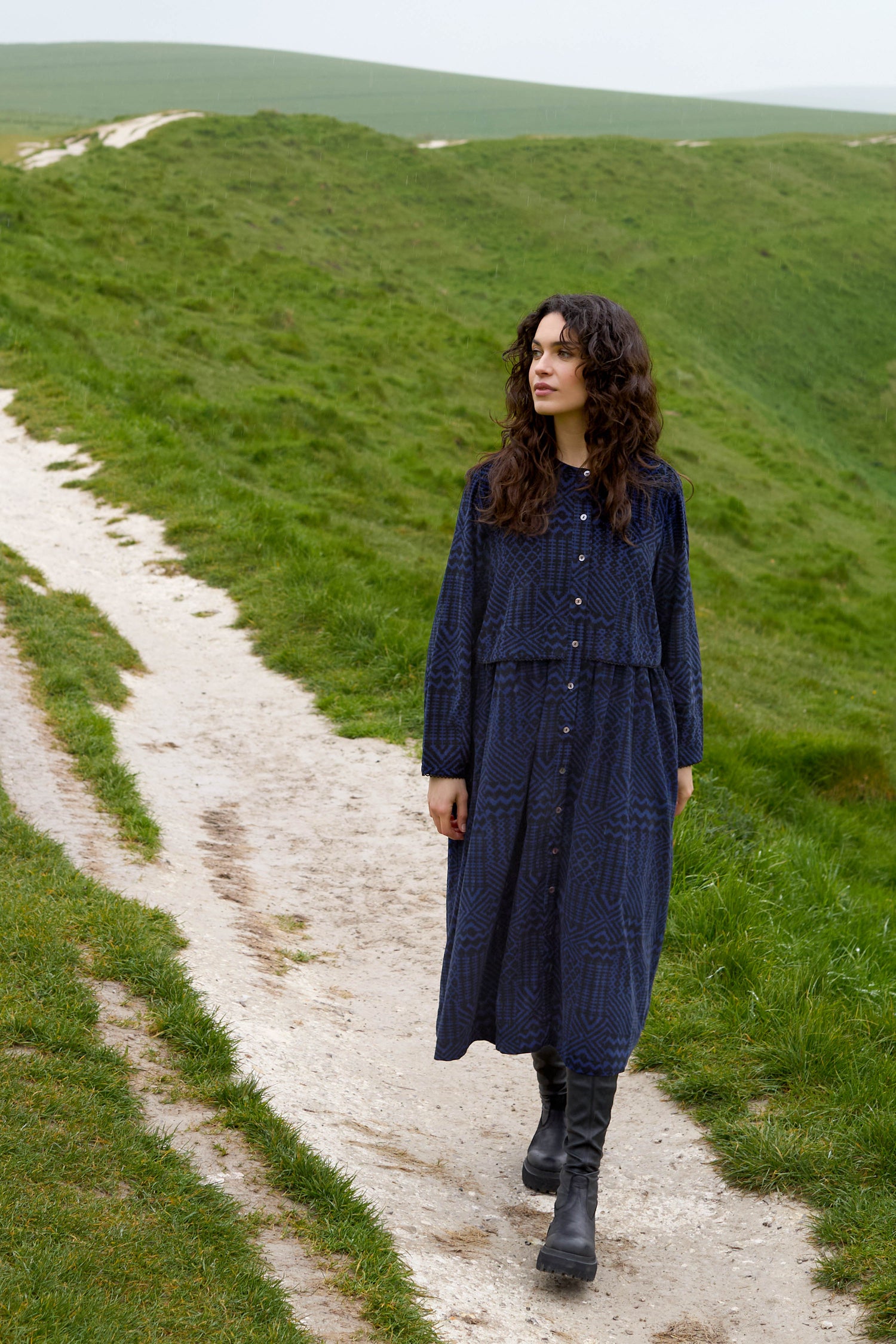 A person in a blue Mono Ikat Printed Cord Dress and black boots strolls along a grassy path.