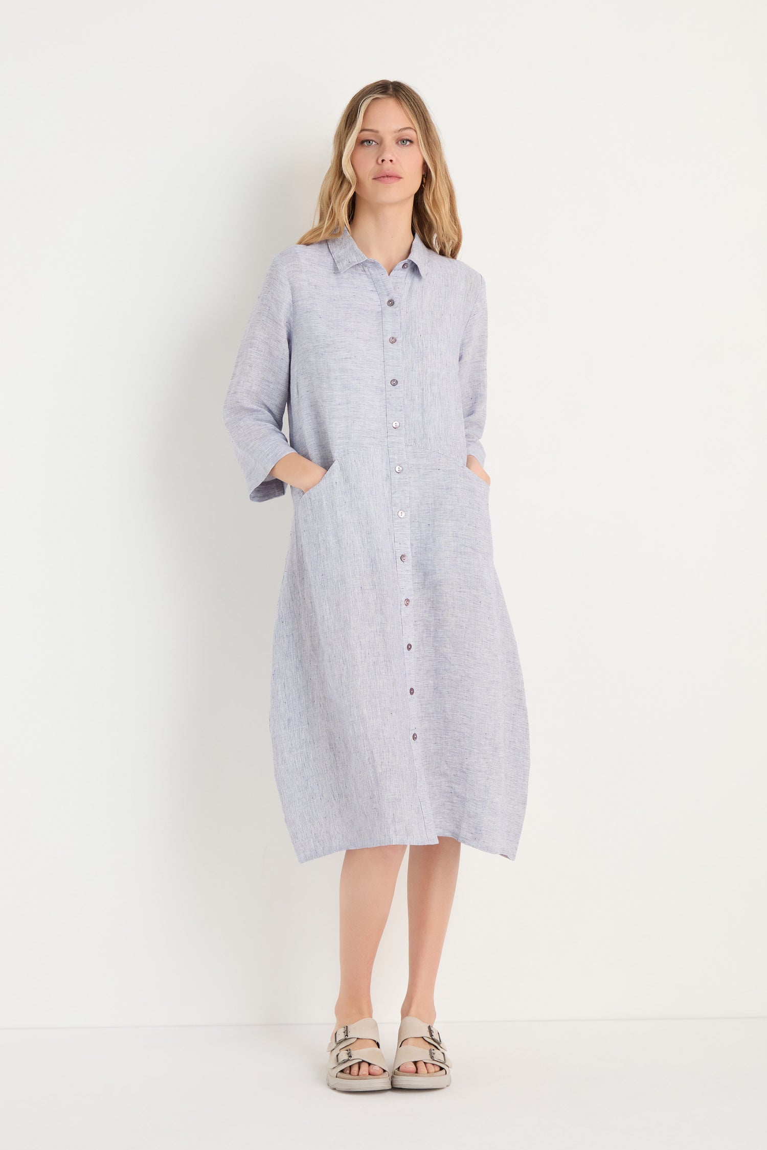 A woman wears the Ticking Linen Stripe Shirt Dress—light blue, long-sleeve, button-down midi with pockets—paired with beige sandals, standing against a plain white background.