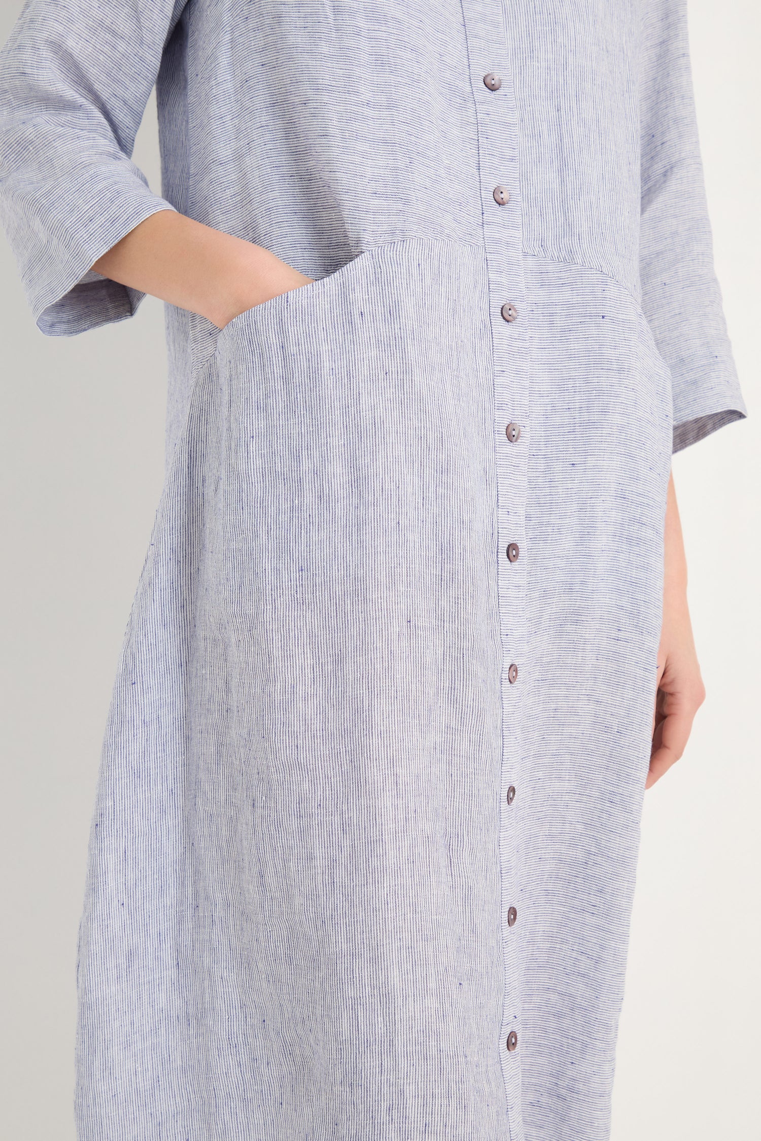 A person wears the Ticking Linen Stripe Shirt Dress in light blue—a relaxed, button-down linen dress with ticking stripes, quarter sleeves, and a large front pocket where their left hand rests.