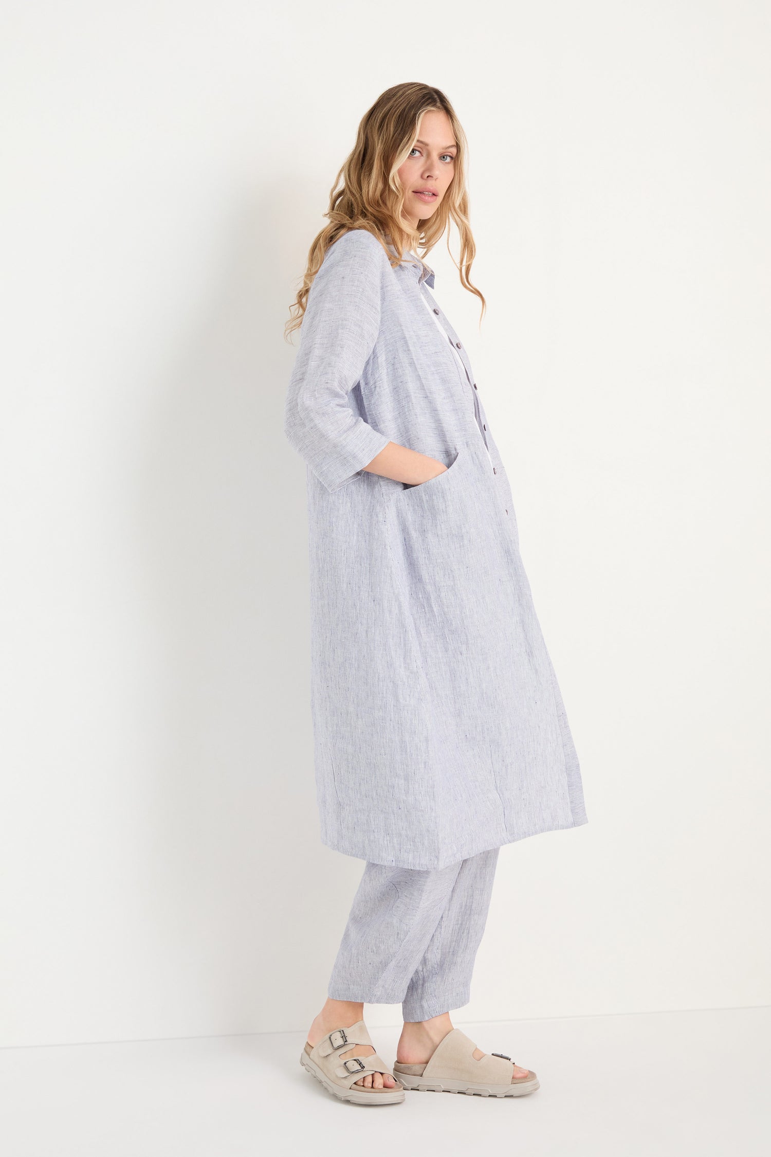 A woman stands sideways in the Ticking Linen Stripe Shirt Dress over matching loose pants and beige sandals against a plain white background.