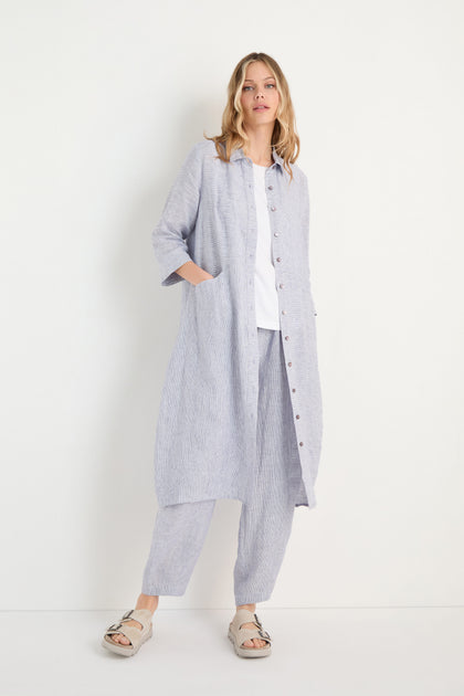 A woman stands against a white background wearing the Ticking Linen Stripe Shirt Dress, paired with matching pants, a white top, and beige sandals.