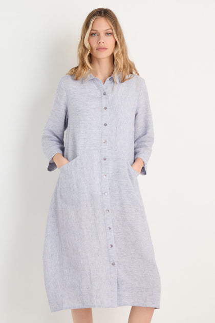 A woman stands against a plain white background wearing the Ticking Linen Stripe Shirt Dress—light blue, relaxed fit, long sleeves, side pockets, and classic linen stripes.