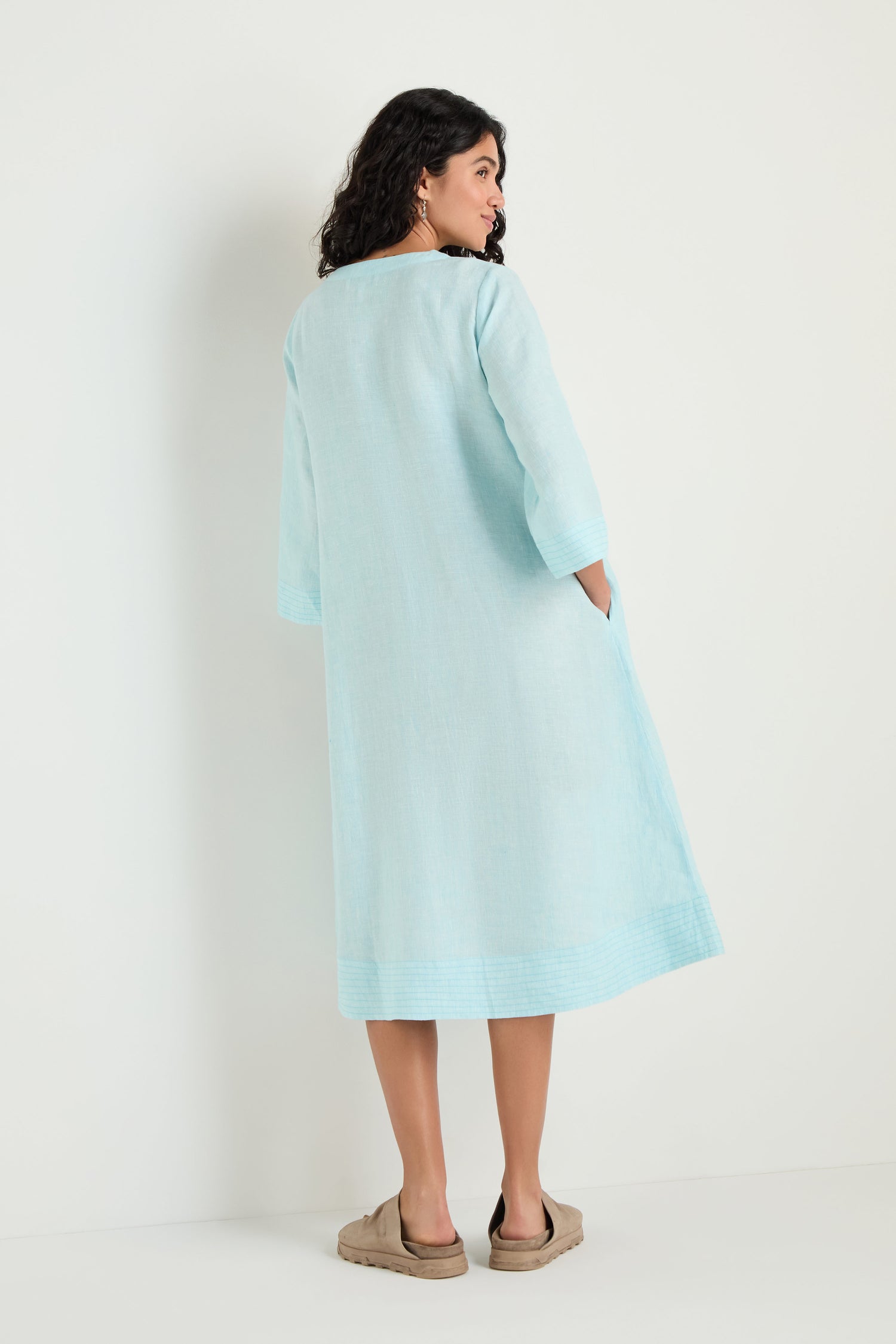 A woman stands against a white background, shown from the back, wearing the Ticking Stripe Linen Button Placket Dress in light blue—a loose, knee-length A-line with three-quarter sleeves—paired with beige slip-on shoes.