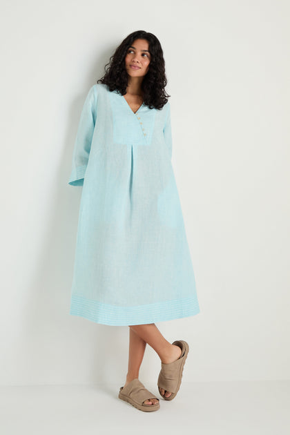 A woman wears the Ticking Stripe Linen Button Placket Dress in light blue with long sleeves and an A-line silhouette, paired with tan sandals, standing against a plain white background.