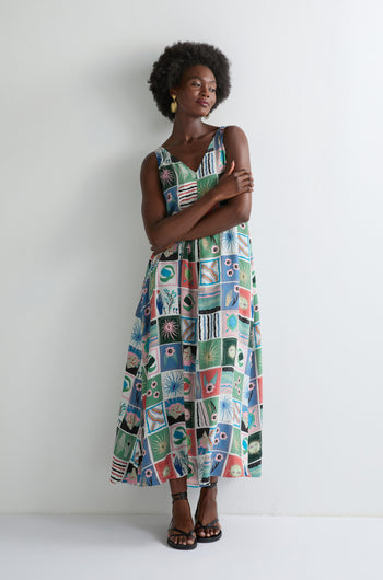 A woman stands against a plain white wall with her arms crossed, wearing the Mystical Collage Sleeveless Dress featuring vibrant geometric and floral patchwork patterns—perfect for summer.