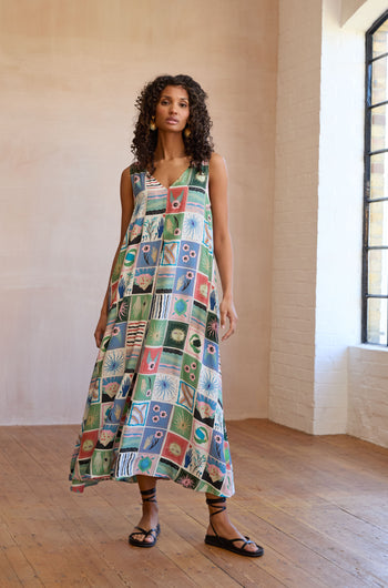 Indoors on wooden floors and against white brick walls, a woman wears the Mystical Collage Sleeveless Dress—a loose, A-line dress with a colorful patchwork pattern—paired with black sandals.