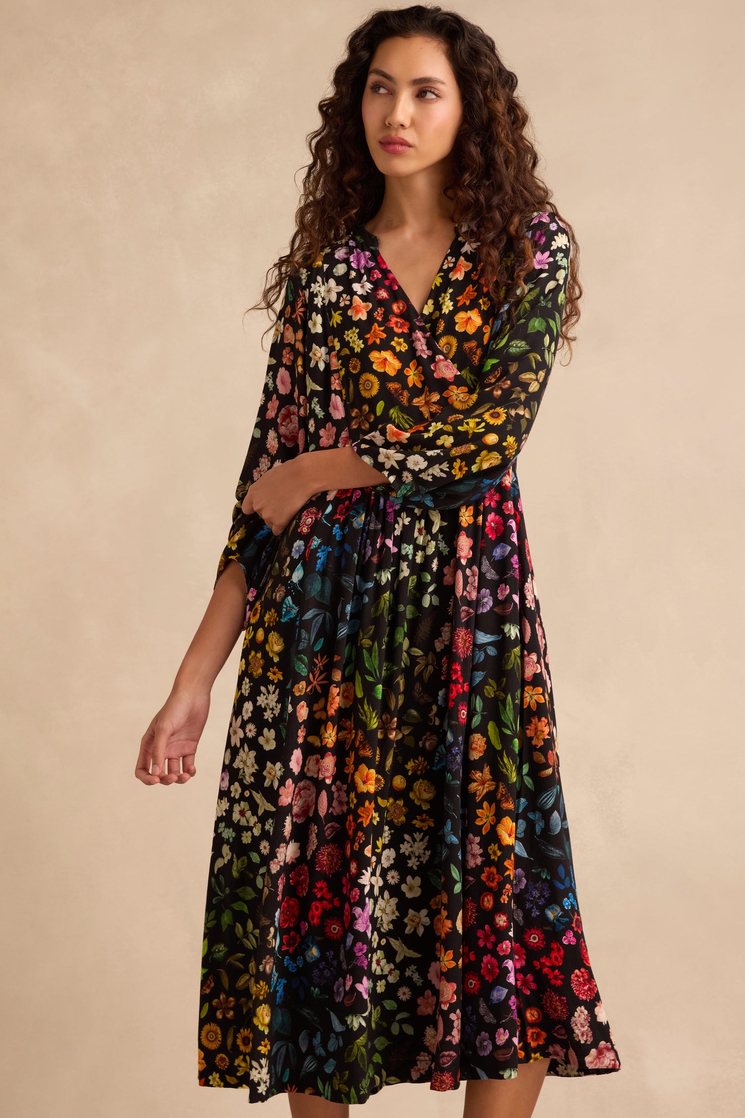 A woman wears the Midnight Garden Viscose Dress, featuring long sleeves, a vibrant floral print on black crepe fabric, and an elegant wrap-front design, as she stands against a beige background.