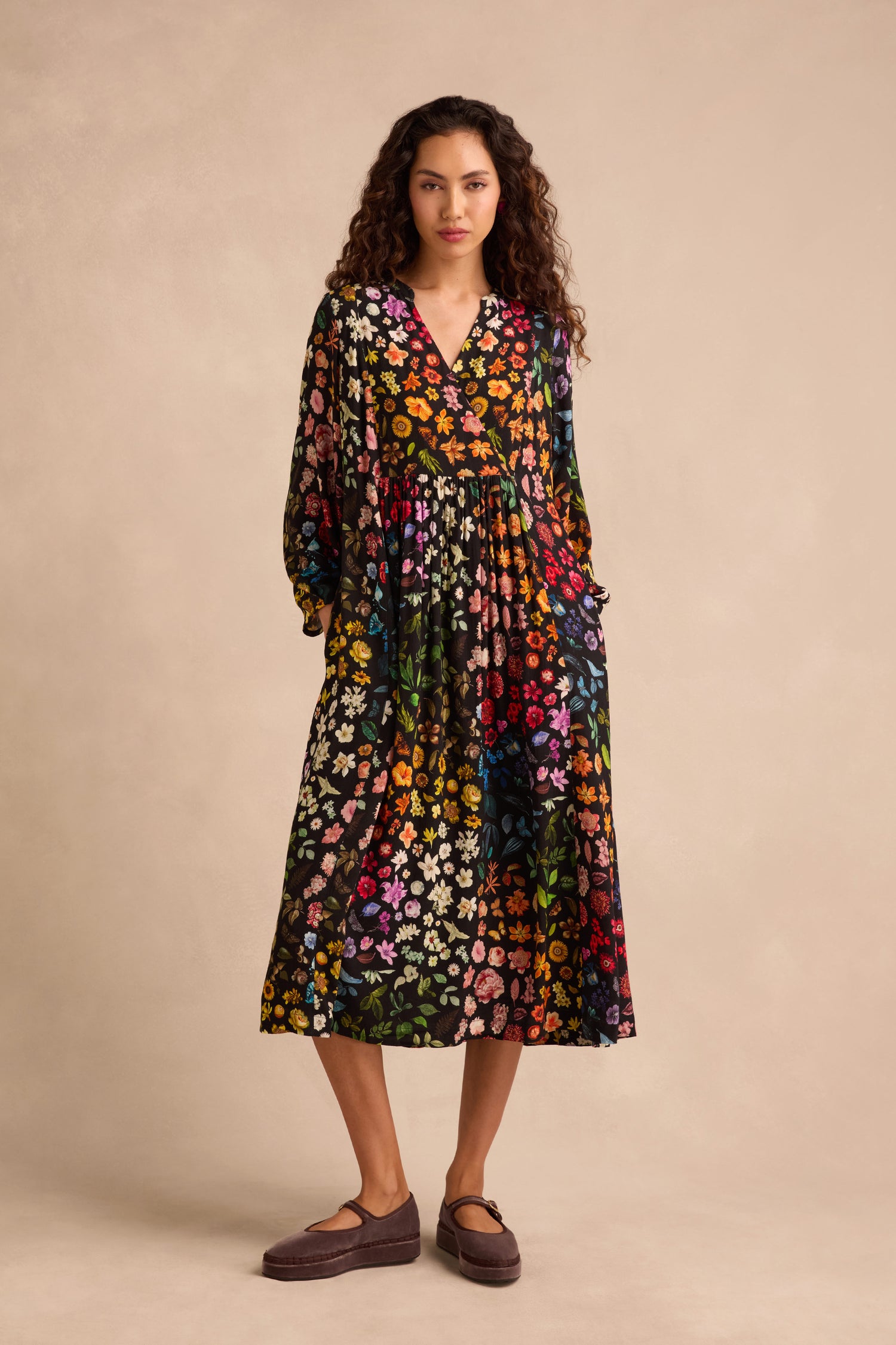 A person stands against a beige background wearing the Midnight Garden Viscose Dress, a long-sleeve, ankle-length black floral crepe dress with a colorful print, paired with brown shoes.