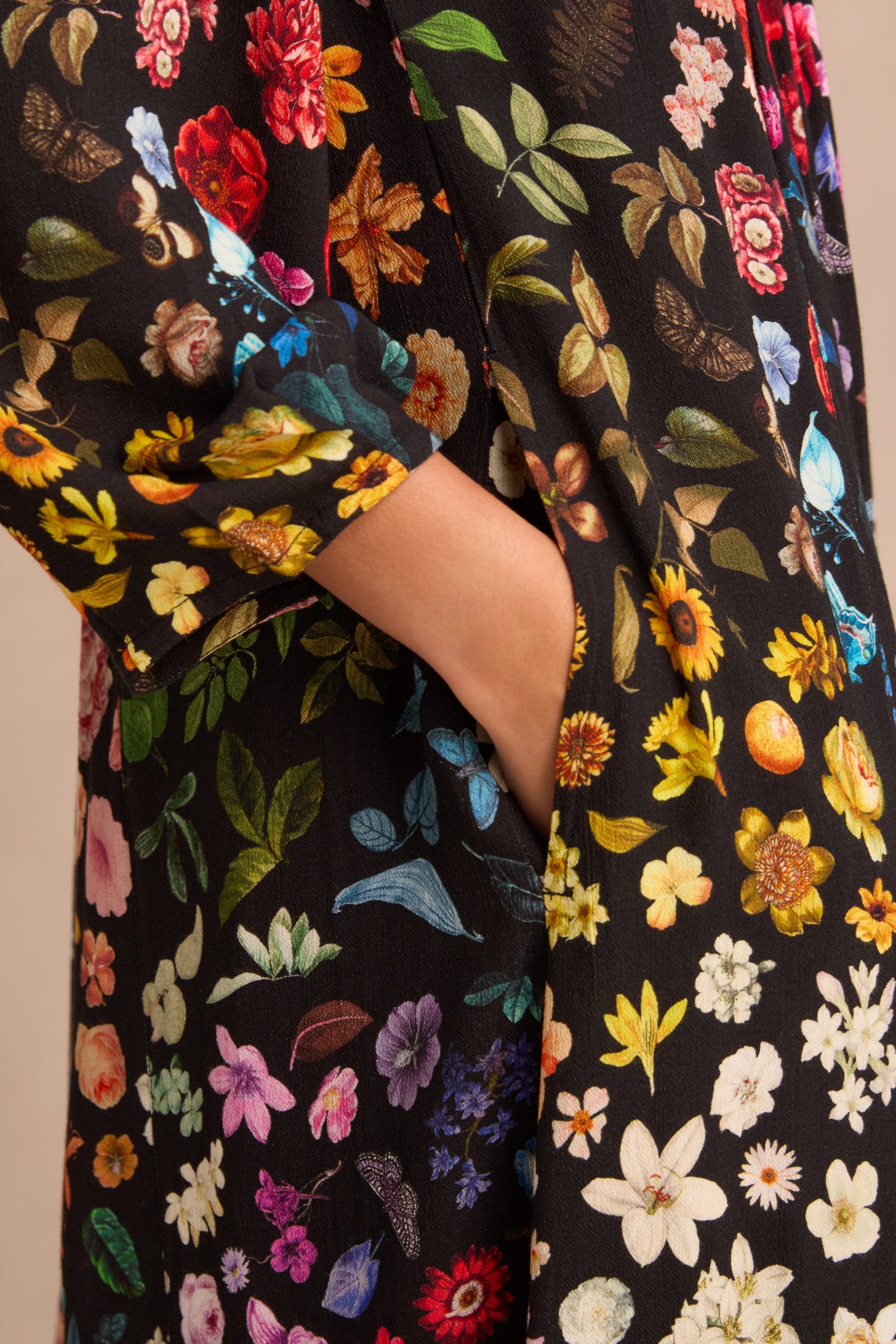 A person’s hand is placed in the pocket of the Midnight Garden Viscose Dress, a black crepe dress adorned with a colorful pattern featuring various types of flowers and leaves.