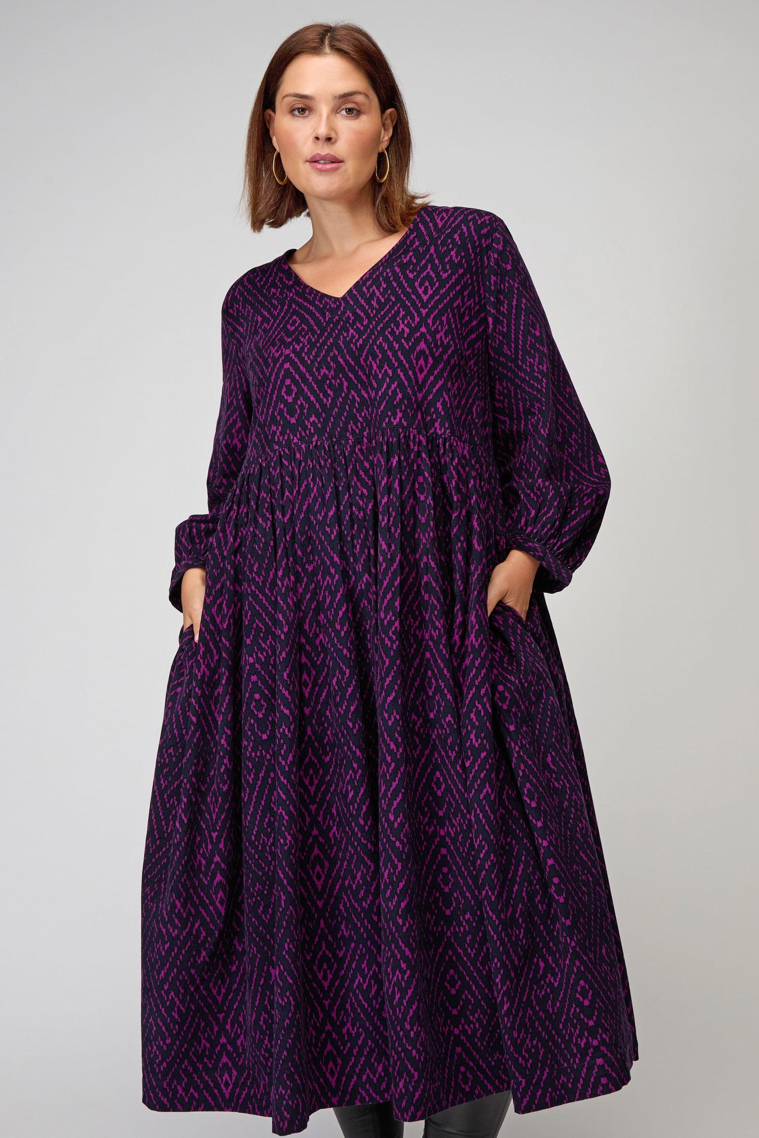 A woman stands against a plain light background wearing the Ornate Textured Plum Cord Dress, featuring long sleeves, a relaxed silhouette, pockets, and rich purple corded fabric.