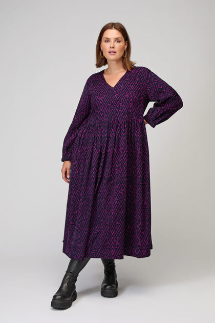 A woman stands against a plain background wearing the Ornate Textured Plum Cord Dress—a long, patterned piece with long sleeves, a relaxed silhouette, and paired with black chunky boots.