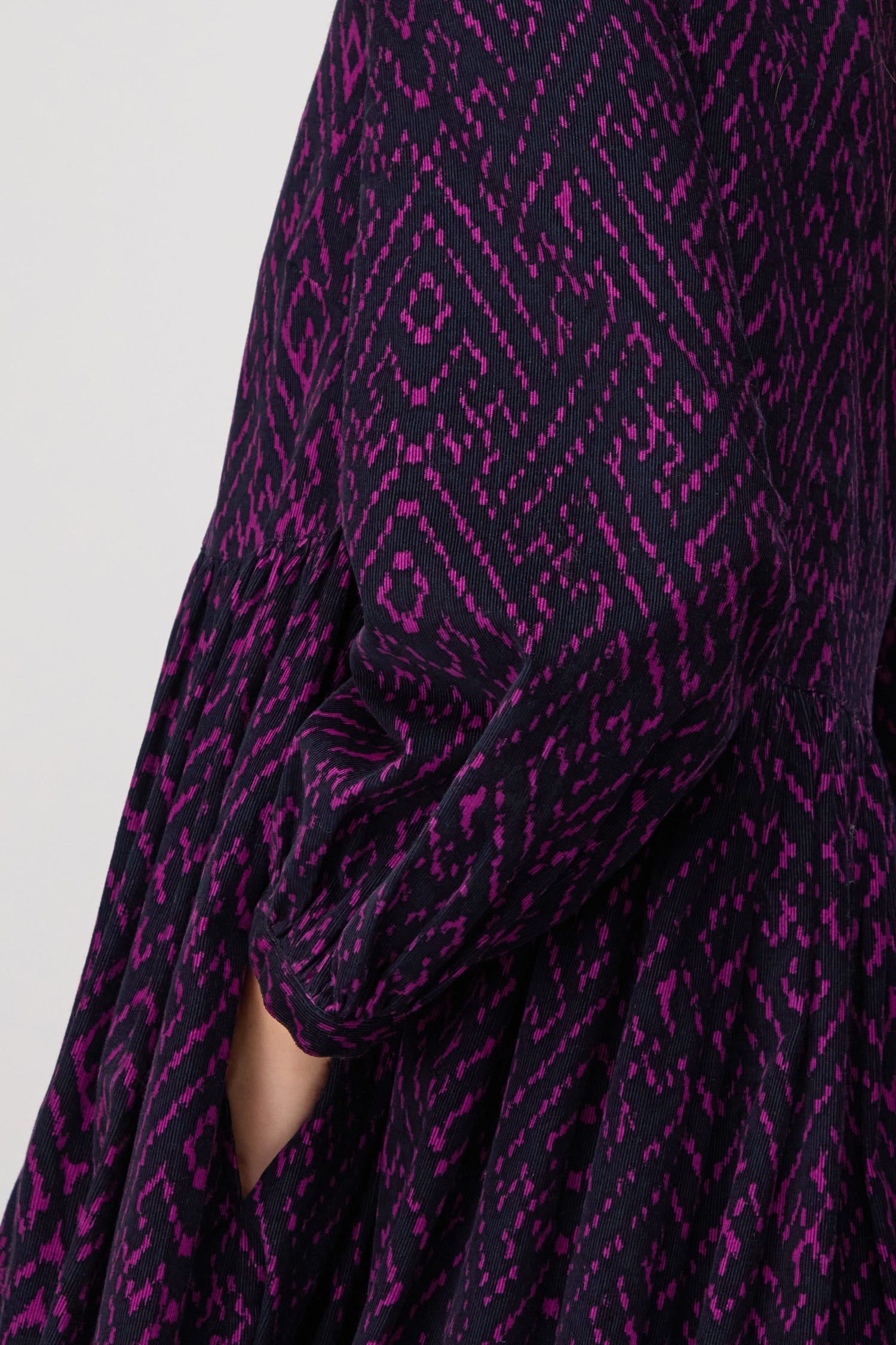 A close-up of the Ornate Textured Plum Cord Dress shows a hand partially tucked into the side pocket, highlighting the relaxed silhouette and rich geometric pattern in dark purple and magenta on the sleeve.