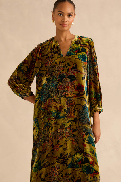 A person wearing a long-sleeved, olive green Silk Velvet Floral Devore Dress in a midi length with a colorful floral and bird pattern.