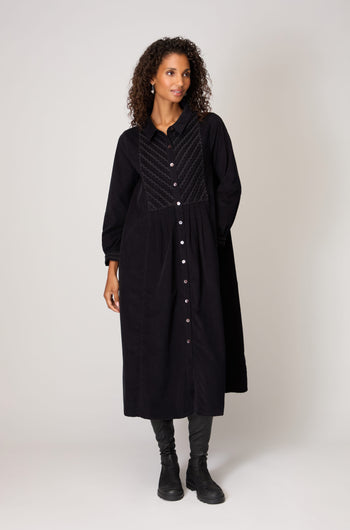 A woman stands against a plain background wearing the Pinwale Cord Ric Rac Detail Dress, a classic fit long black button-down cord dress with textured front panel, paired with black pants and boots.