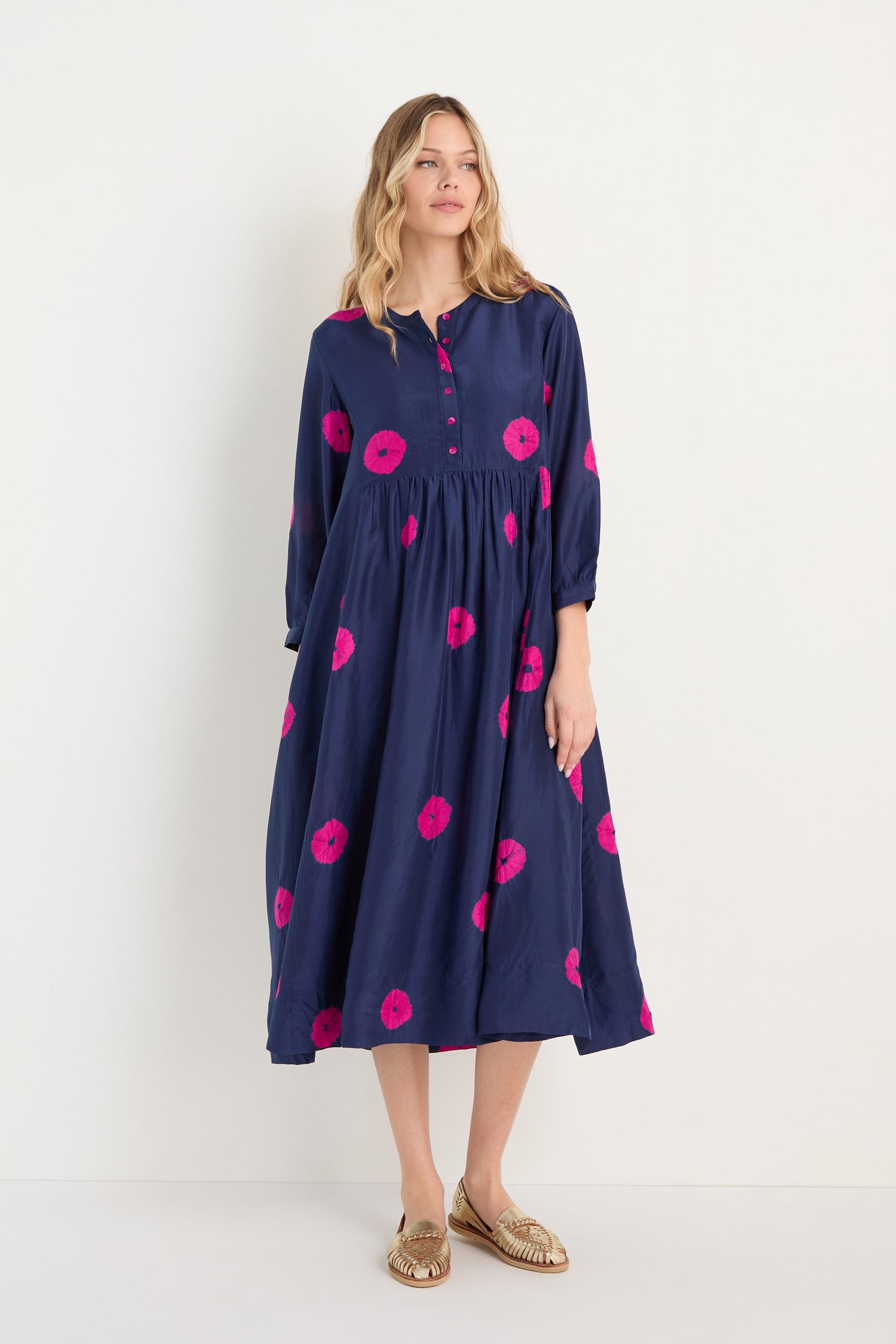 A woman models the Shibori Spot Silk Dress, featuring navy blue silk with pink circular patterns and long sleeves, paired with gold flats, set against a plain white background.