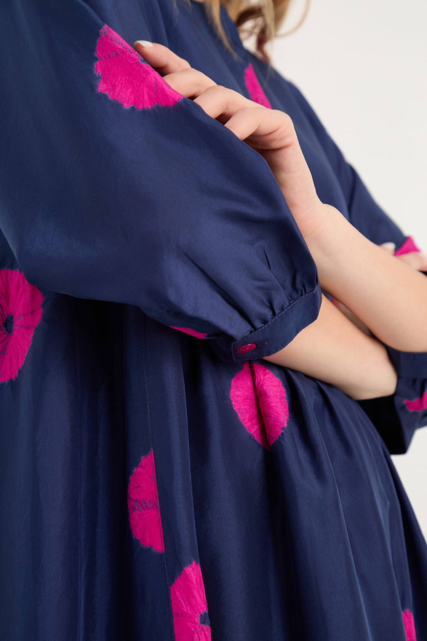 Model wearing the Shibori Spot Silk Dress in navy blue with bright pink circular patterns, shown from neck to mid-torso with a bent arm and hand near her face, highlights the dress's contemporary silhouette.