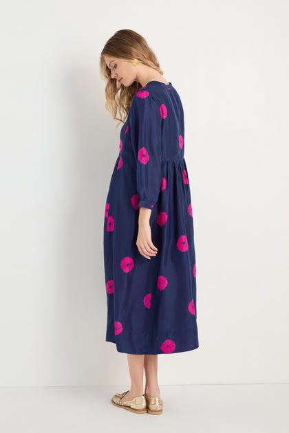 A woman stands sideways against a white background wearing the Shibori Spot Silk Dress—navy blue with pink circular patterns and a modern silhouette—paired with gold flat sandals.