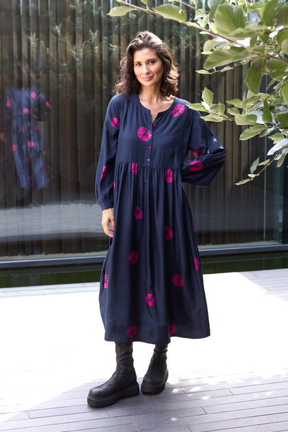 A woman stands outdoors in the Shibori Spot Silk Dress—a modern navy blue silk dress with pink shibori spots—paired with black boots, against a backdrop of plants and a glass wall.