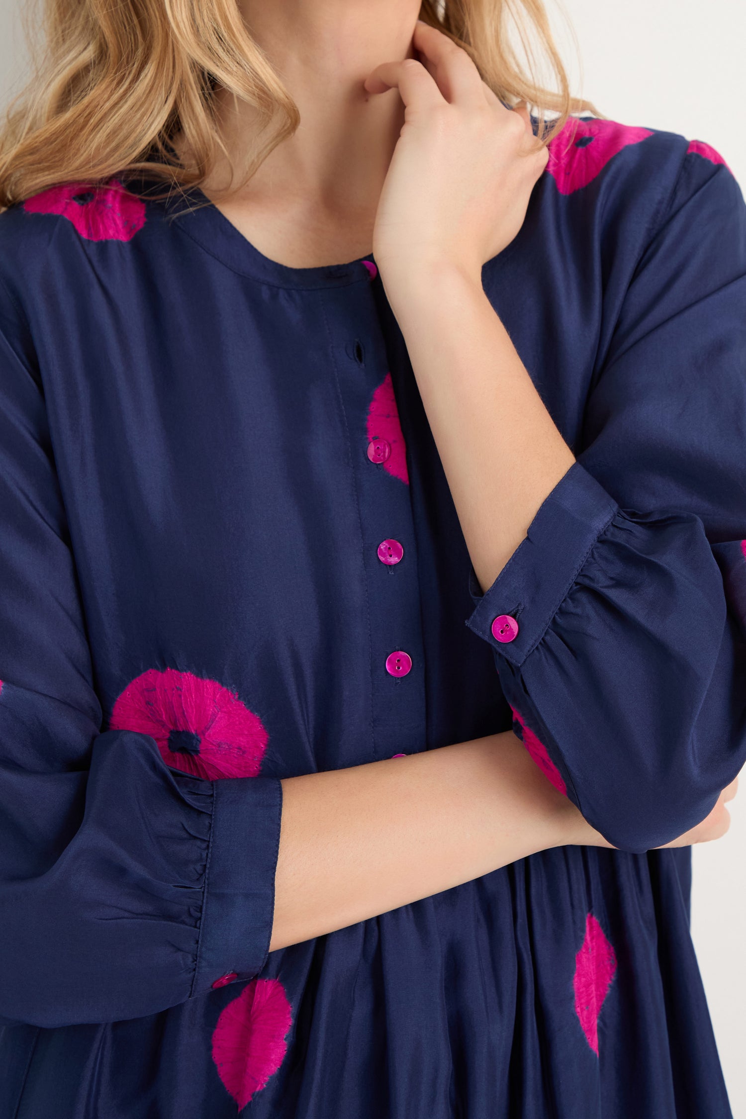Woman in a Shibori Spot Silk Dress—navy blue with bright pink circular patterns—stands with one hand touching her neck and the other arm crossed.