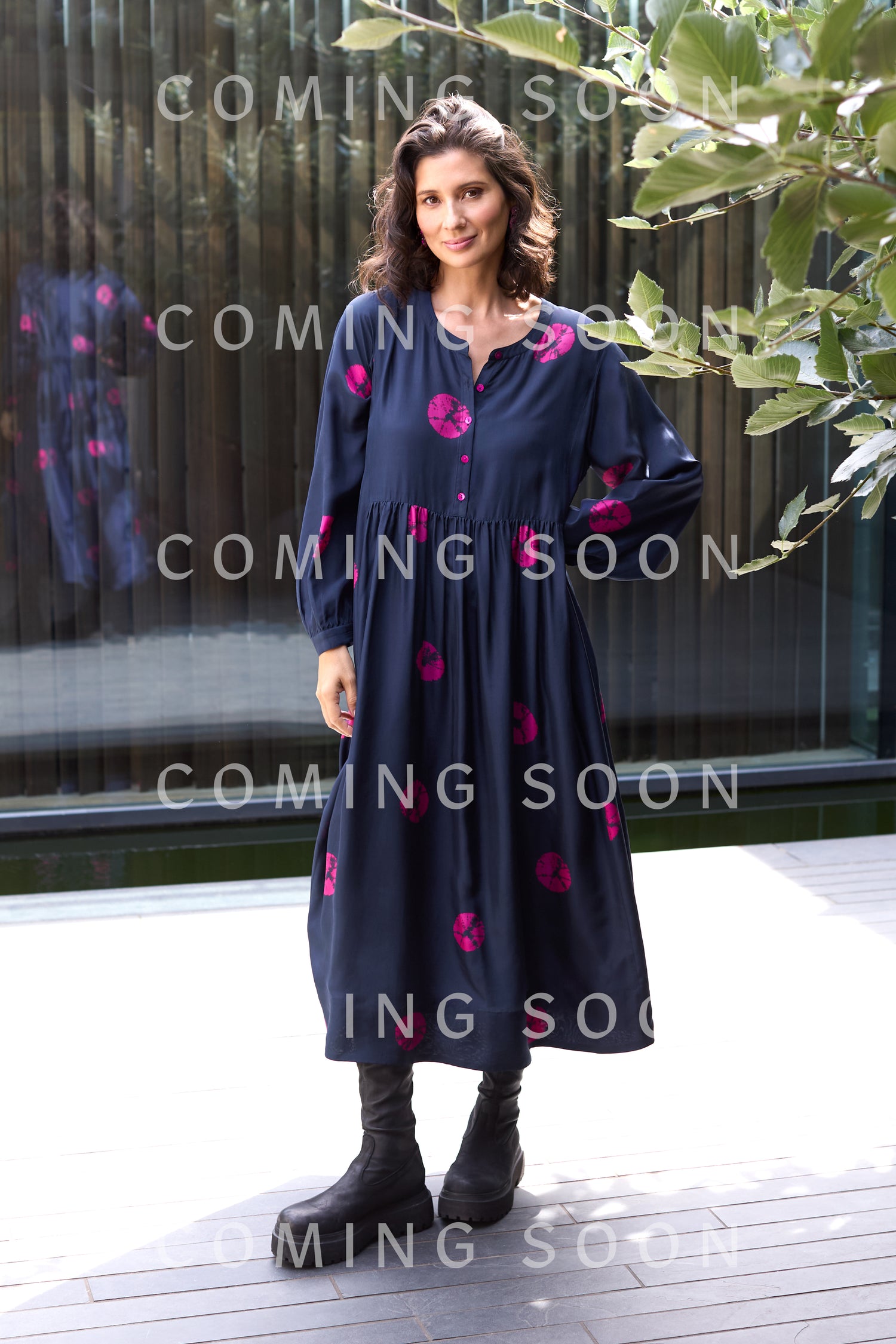 A woman stands outdoors in the Shibori Spot Silk Dress—long, navy with pink patterns—paired with black boots and a modern silhouette. "Coming Soon" appears repeatedly over the image.