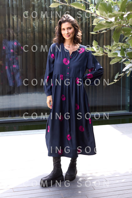 A woman stands outdoors in the Shibori Spot Silk Dress—long, navy with pink patterns—paired with black boots and a modern silhouette. "Coming Soon" appears repeatedly over the image.