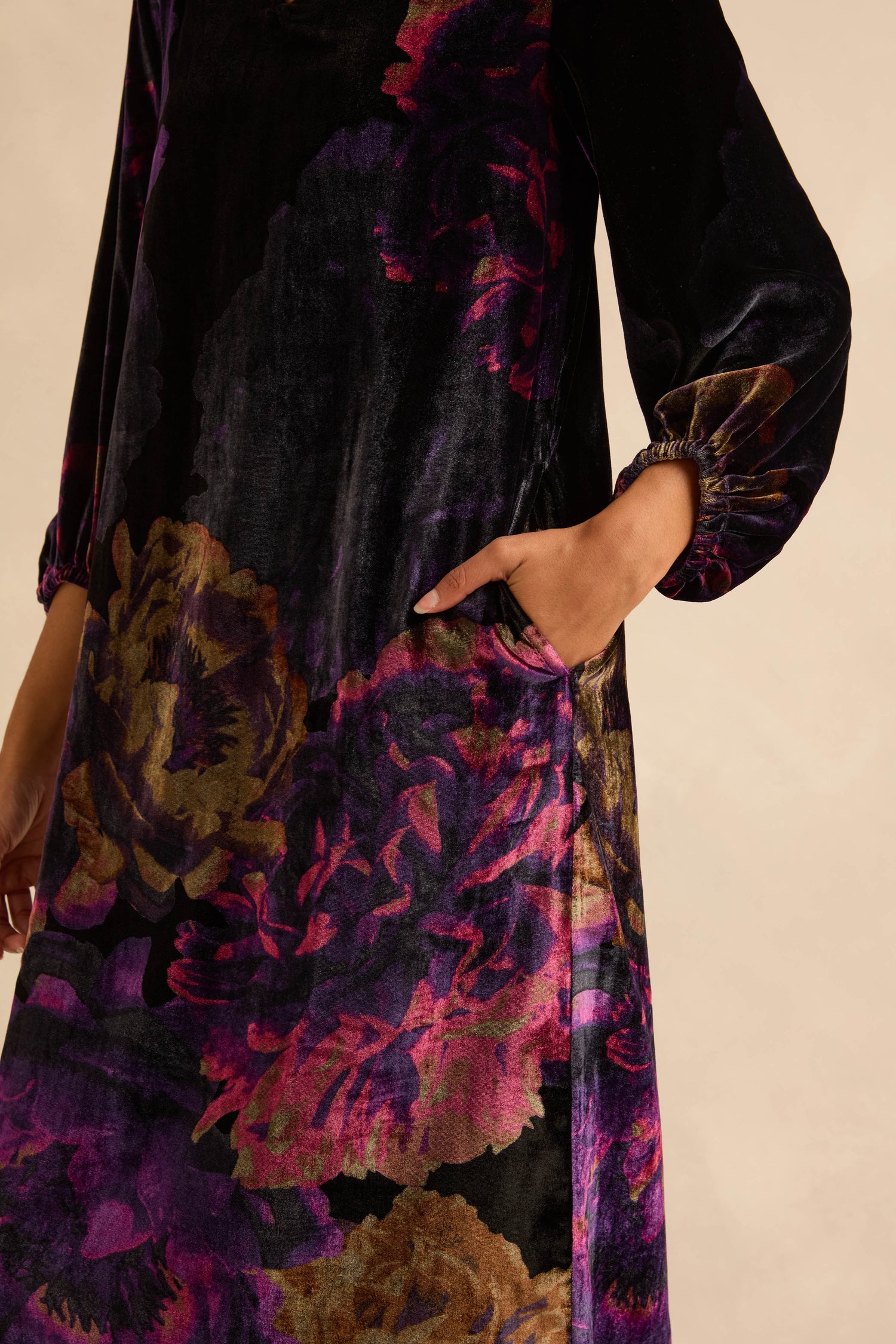 The Midnight Peonies Velvet Dress is a long-sleeved, midi-length velvet dress with a black background and large purple and bronze photographic floral prints, shown with a hand in the pocket.